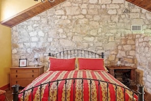 A bedroom with a stone wall, a metal bed frame, and a wooden nightstand. - Fred Haus - Historic home with Hot Tub & Fire Pit (Fredericksburg)
