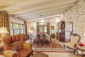 A spacious room with a stone wall, a wooden ceiling, a sofa, a dining table with chairs, and a chandelier. - Fred Haus - Historic home with Hot Tub & Fire Pit (Fredericksburg)