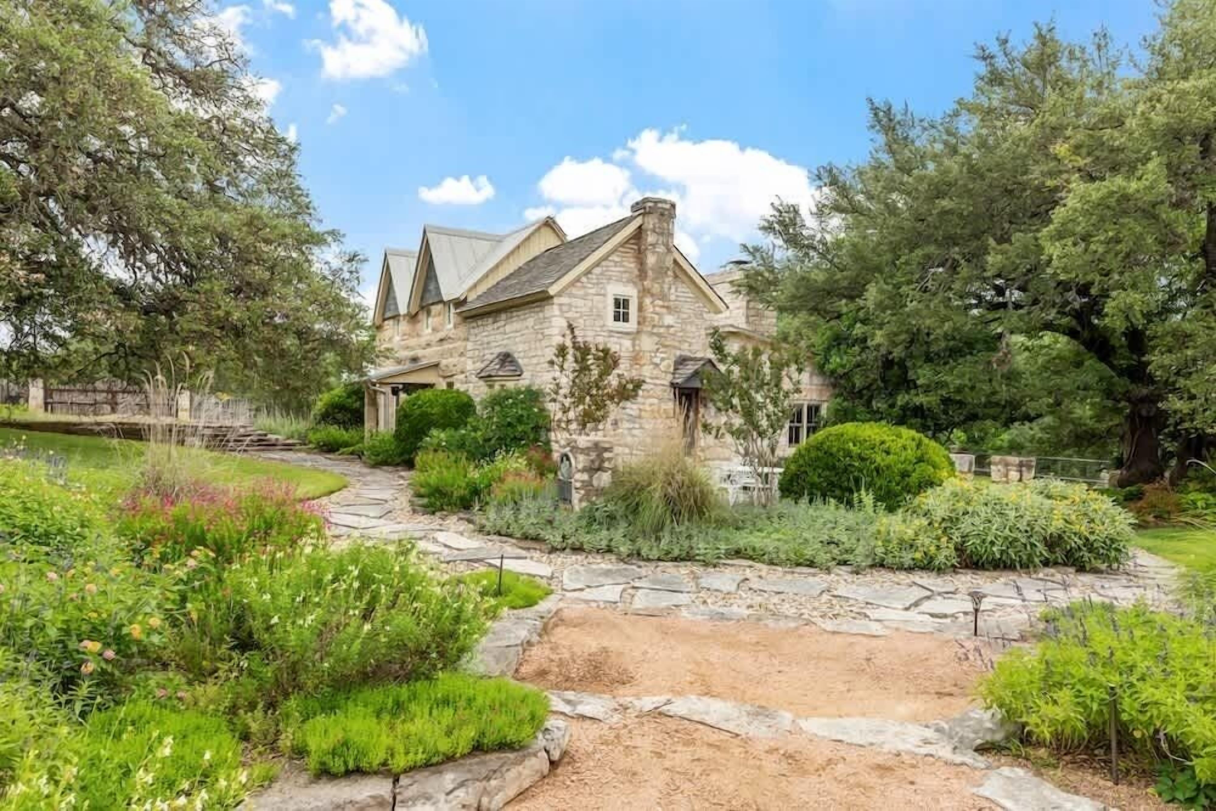 A stone house with a stone pathway leading to the entrance, surrounded by lush greenery and trees.