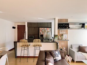 A modern living space with a kitchen, two bar stools, a sofa, and a bookshelf. - 2-bedroom apartment in beautiful El Poblado Medellín with AC (Medellín)