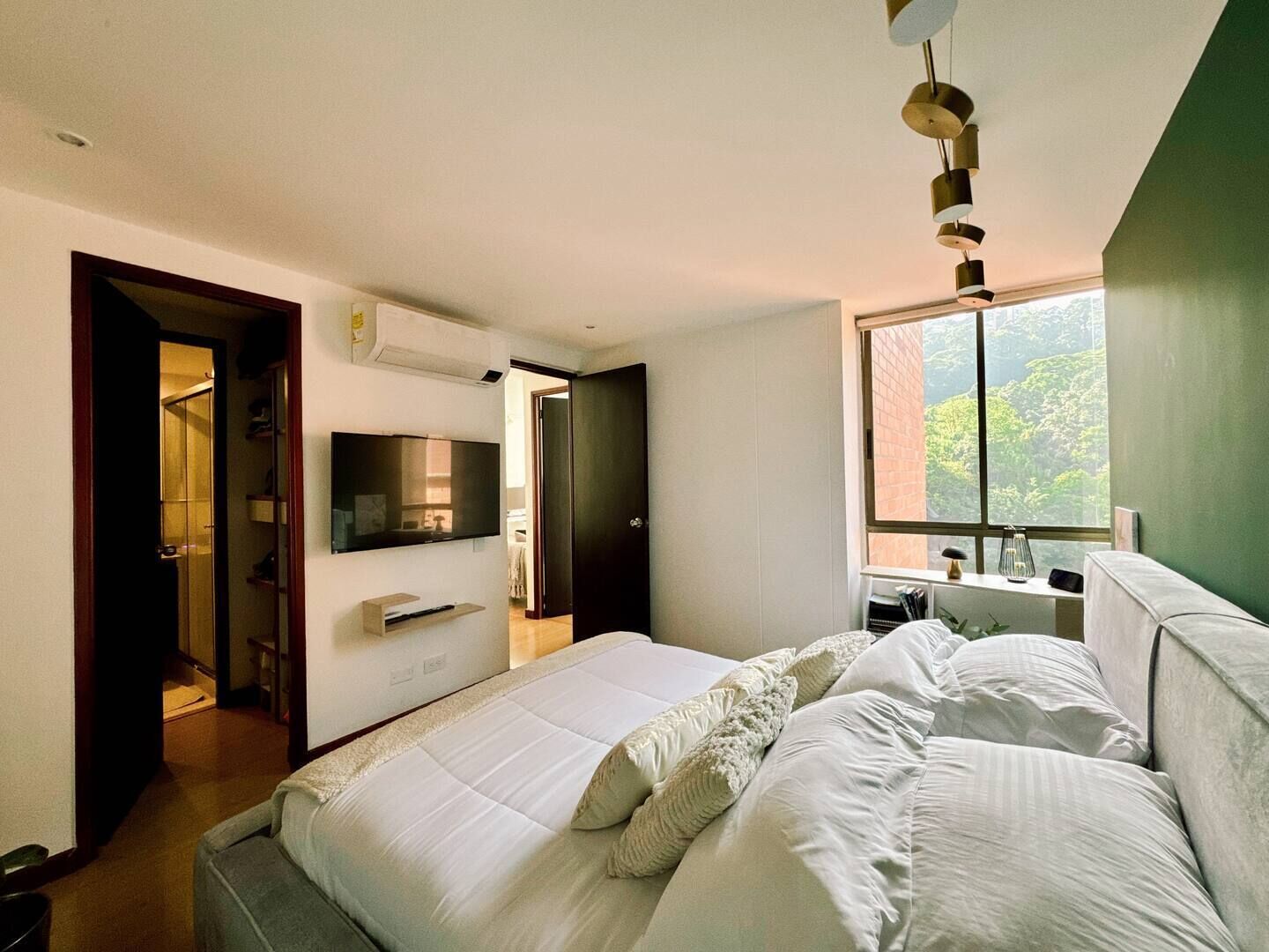 A modern bedroom with a large bed, a TV mounted on the wall, and a window providing a view of greenery outside.