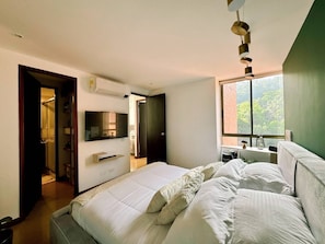 A modern bedroom with a large bed, a TV mounted on the wall, and a window providing a view of greenery outside. - 2-bedroom apartment in beautiful El Poblado Medellín with AC (Medellín)