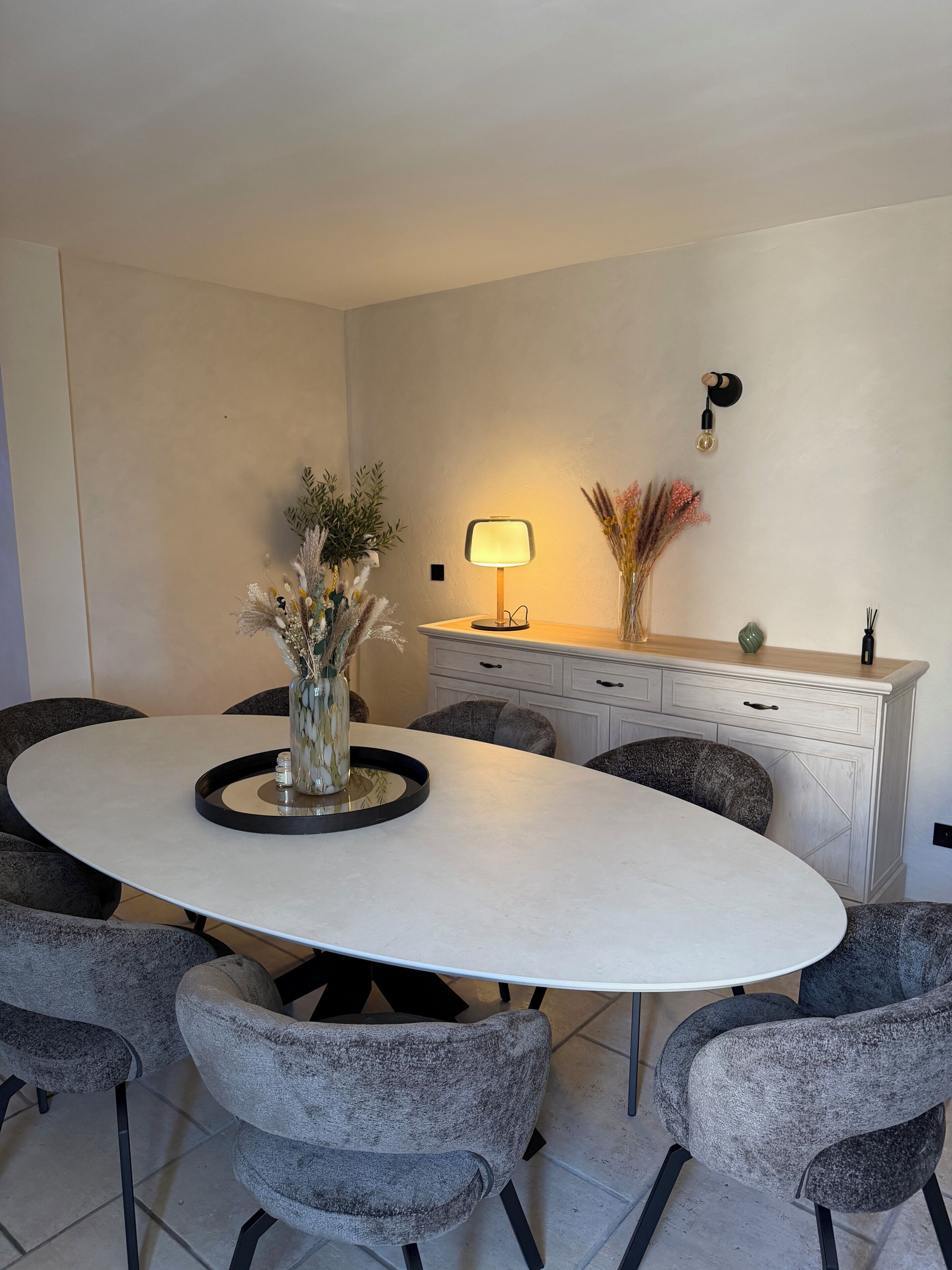A dining area with a round table, grey upholstered chairs, a vase with dried flowers, a wall-mounted lamp, and a white cabinet.
