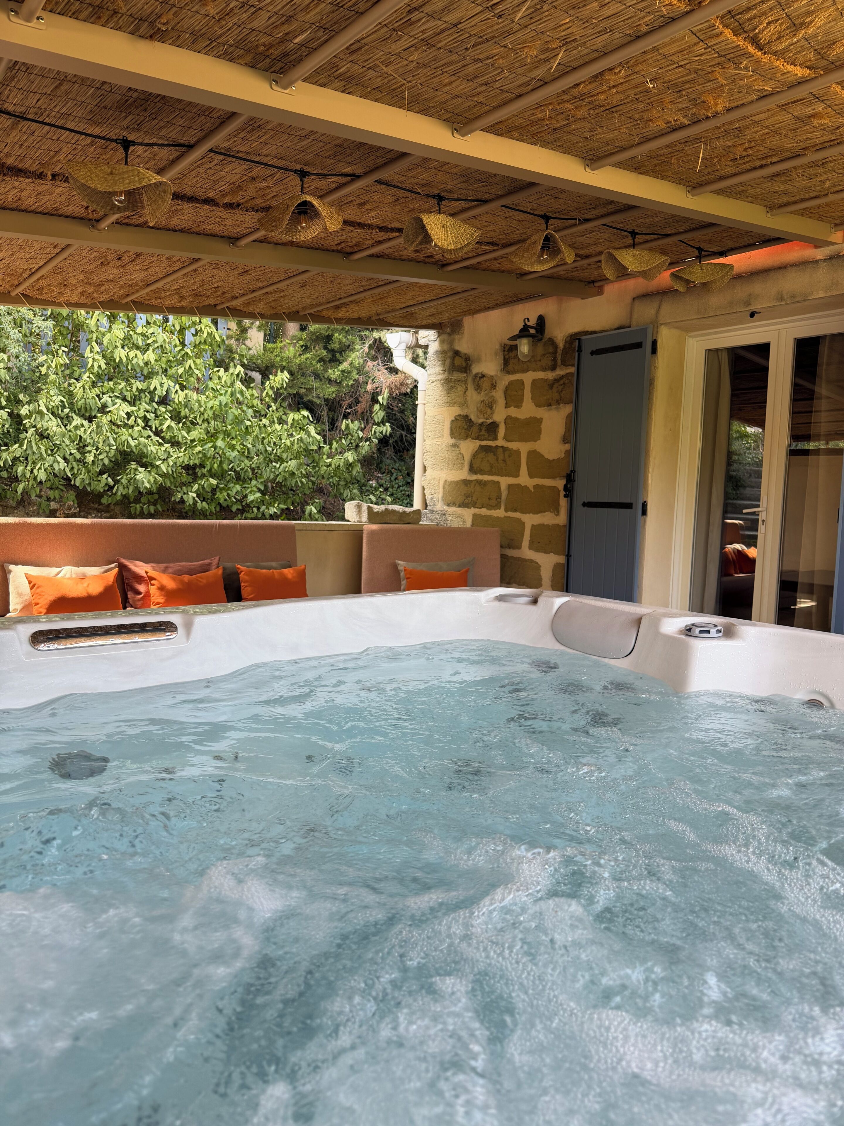 A hot tub with a stone wall and orange cushions, a thatched roof, and a view of greenery.