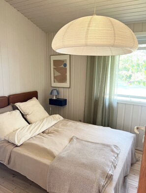 A bedroom with a bed, a bedside table with a lamp, a hanging light fixture, a framed picture on the wall, and a window with curtains. - Classic Danish summer house (Asnæs)