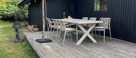 A wooden deck with a table and chairs, a pergola with an umbrella, and a dark building in the background.