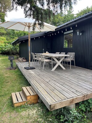 A wooden deck with a table and chairs, a pergola with an umbrella, and a dark building in the background. - Classic Danish summer house (Asnæs)