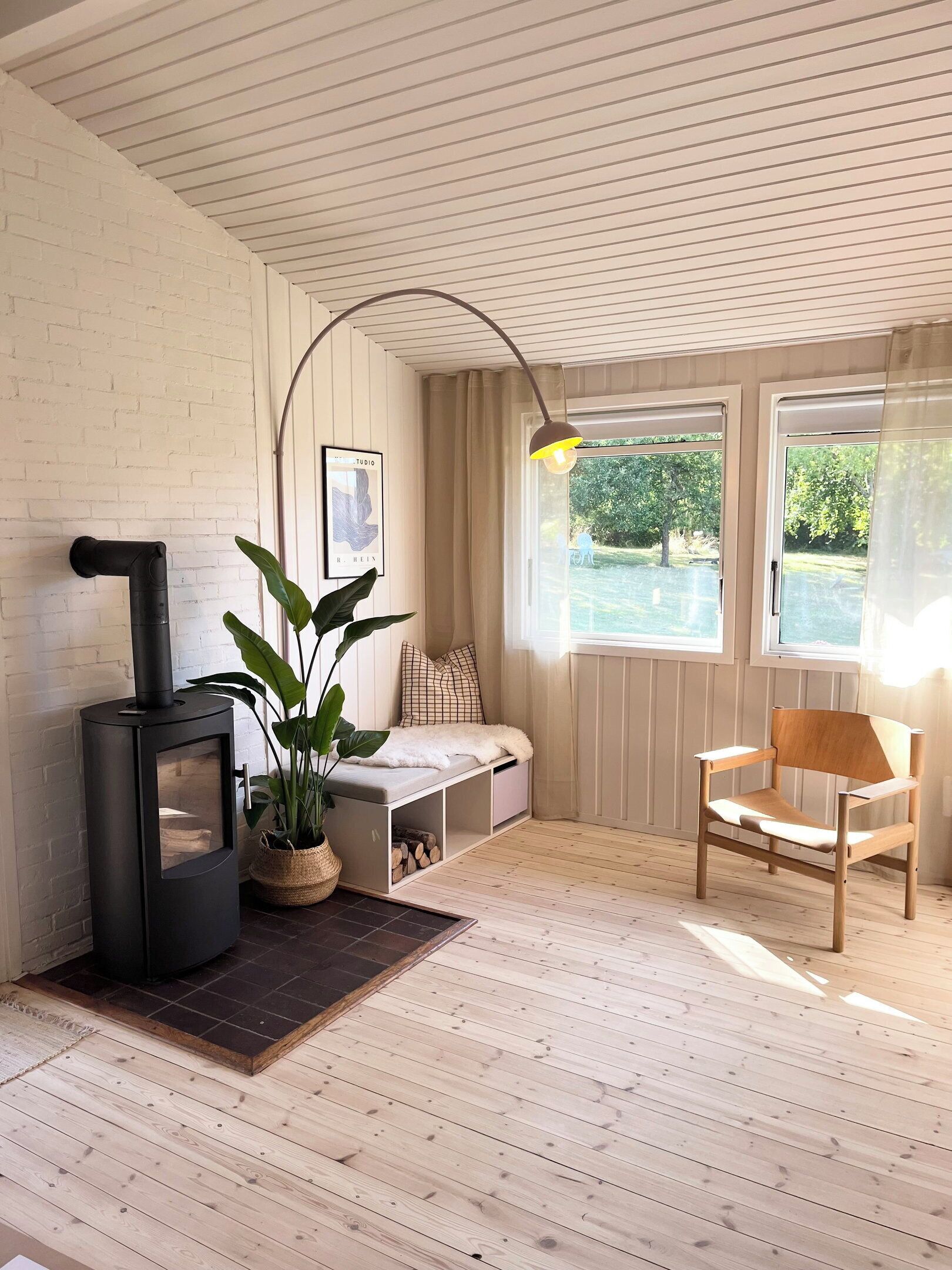A cozy room with a wood-burning stove, a bench, a chair, and a large window with a view of greenery.