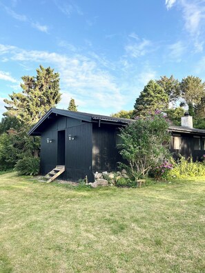 A black wooden shed with a door and a small set of wooden steps leading up to it, surrounded by grass and various plants. - Classic Danish summer house (Asnæs)