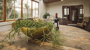 Une table à manger sur laquelle se trouve un grand saladier vert rempli d’herbes fraîches, une bouteille de vin, et qui offre une vue sur un coin repas avec des chaises et une fenêtre.