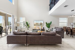 A modern living room with a sectional sofa, a dining area with wooden chairs, and a large window with blinds. - ViLla dReaMs PoOl +Game Room NeAr DiSnEy (Kissimmee)