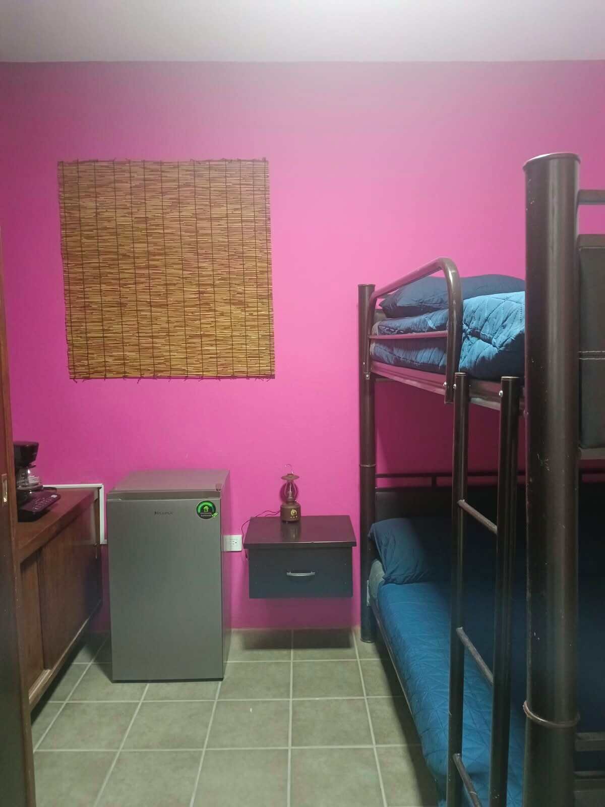 A compact room with a bunk bed, a desk with a computer, a small refrigerator, and a window with a bamboo blind.