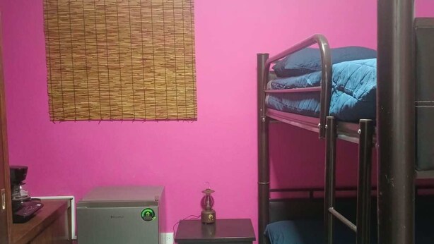 Een kleine kamer met een stapelbed, een bureau met een computer, een kleine koelkast en een raam met een bamboe rolgordijn.