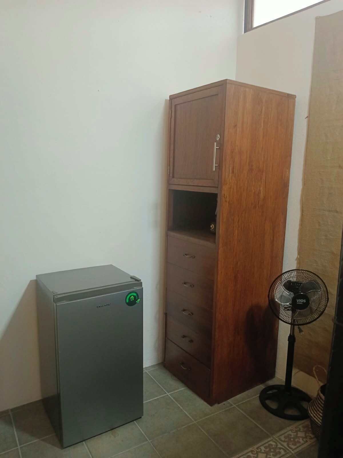 Una habitación pequeña con un electrodoméstico metálico, un armario de madera y un ventilador de pie.