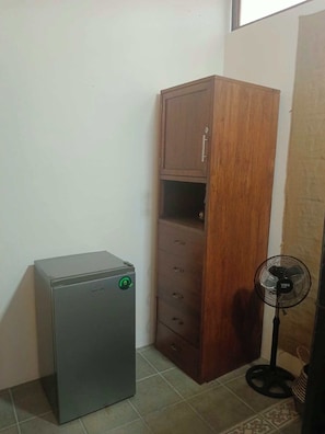 Una habitación pequeña con un electrodoméstico metálico, un armario de madera y un ventilador de pie.