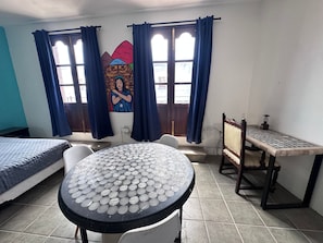 Una habitación con una cama, una mesa con tablero estampado, una silla, un escritorio con lámpara y una ventana con cortinas.