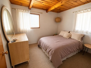 A bedroom with a wooden ceiling, a bed with a pink bedspread, a round wall hanging, and a mirror on a wooden dresser.