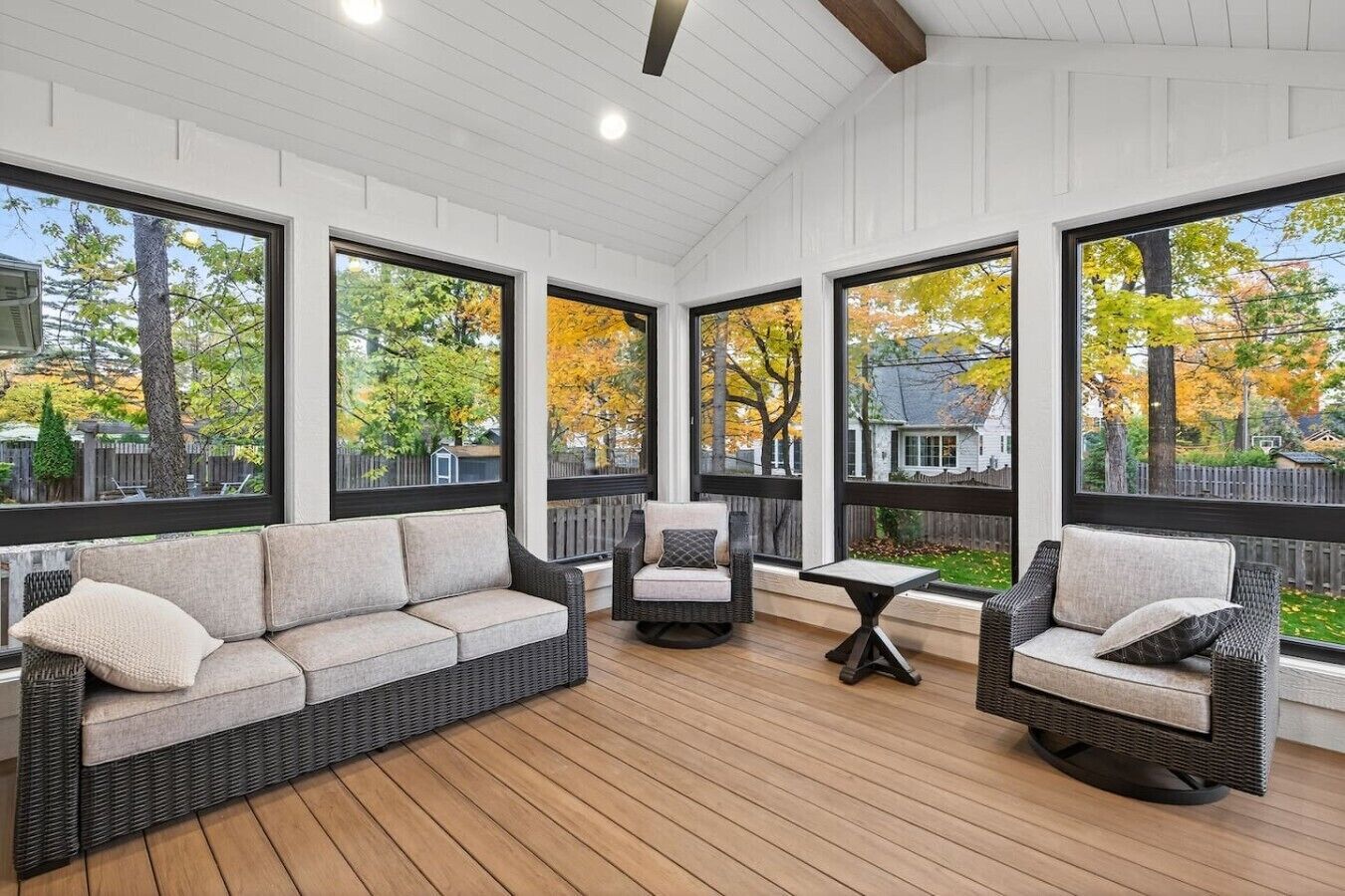 Un solarium con suelo de madera, mobiliario de mimbre y ventanas amplias con vista a un barrio residencial.