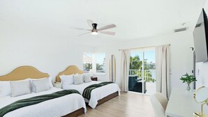 Ein Hotelzimmer mit zwei Betten, einem Schreibtisch, einem Stuhl, einem Deckenventilator und einem Balkon mit Vorhängen.