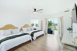 Ein Hotelzimmer mit zwei Betten, einem Schreibtisch, einem Stuhl, einem Deckenventilator und einem Balkon mit Vorhängen.