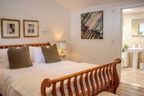 Habitación de hotel con cama, cabecera de madera, mesita de noche con lámpara, un cuadro en la pared y un baño visible a través de una puerta abierta.