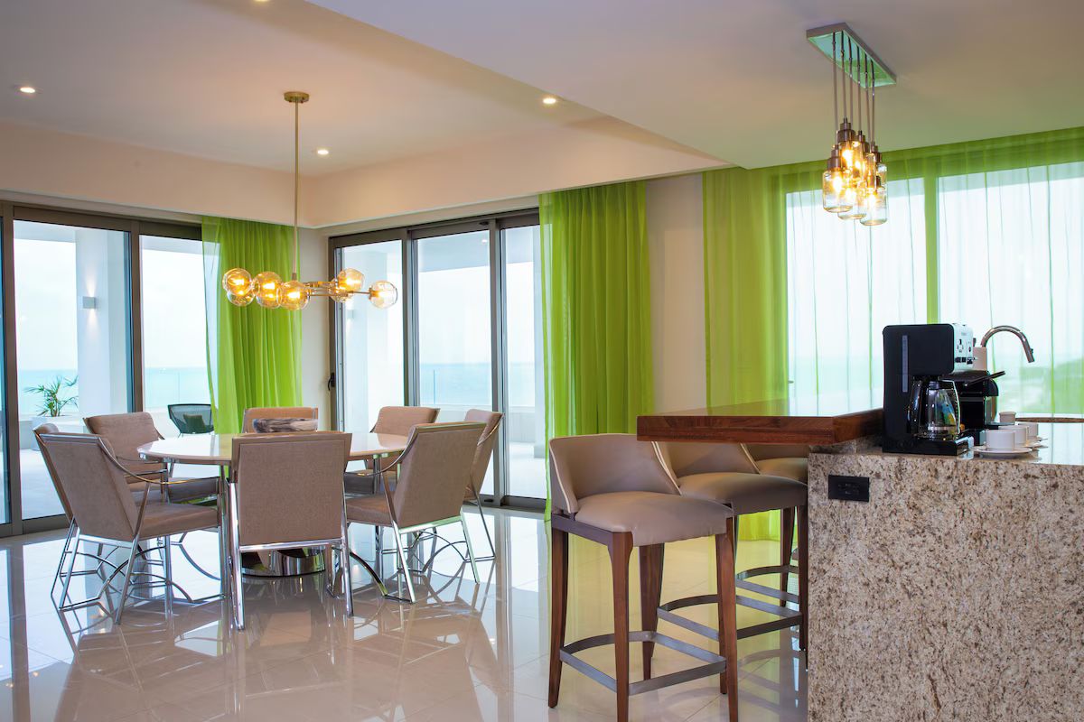 A modern hotel room with a dining area, a kitchen counter, and large windows with green curtains.