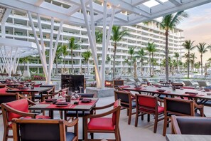 A hotel dining area with multiple tables and chairs, a pool in the background, and a modern building facade. - Garza Blanca Luxurious Oceanfront 3-Bedroom with Expansive Balcony and Jacuzzi (Cancún)