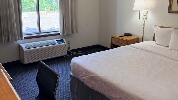 Habitación de hotel con cama, escritorio, silla, lámpara de noche y ventana con cortinas.