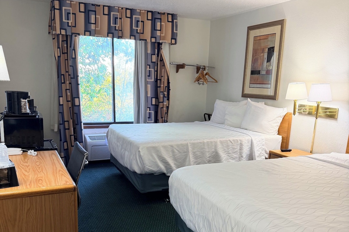 A hotel room with two beds, a desk with a coffee maker, a trash bin, and a window with curtains.