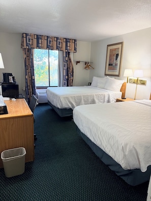 Habitación de hotel con dos camas, un escritorio con cafetera, un cesto de basura y una ventana con cortinas.