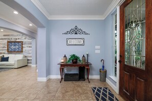 A hallway with a welcome sign, a console table with decorative items, a patterned rug, and a wooden door with a glass panel. - Large Farm House Theme Home on Wooded Lot and Still Close to Everything (Spring)