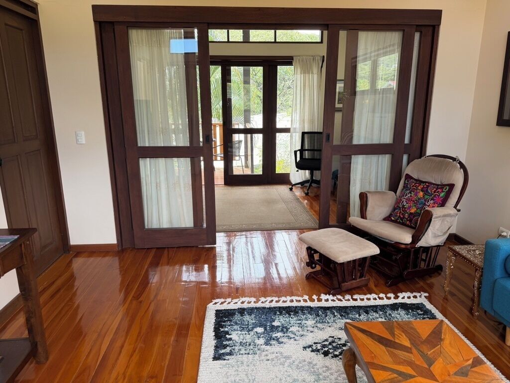 An interior with a wooden floor, a sofa, a chair, and a view through a glass door to an outdoor area.