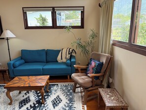 Ein Wohnzimmer mit einem blauen Sofa, einem Holzsesseln, einem Couchtisch mit Muster, einer Stehlampe und Blick auf Grün hinter den Fenstern.