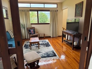 Ein Zimmer mit Holzboden, einer Couch, einem Sessel, einem kleinen Tisch, einem Fernseher und einem Fenster mit Vorhängen.