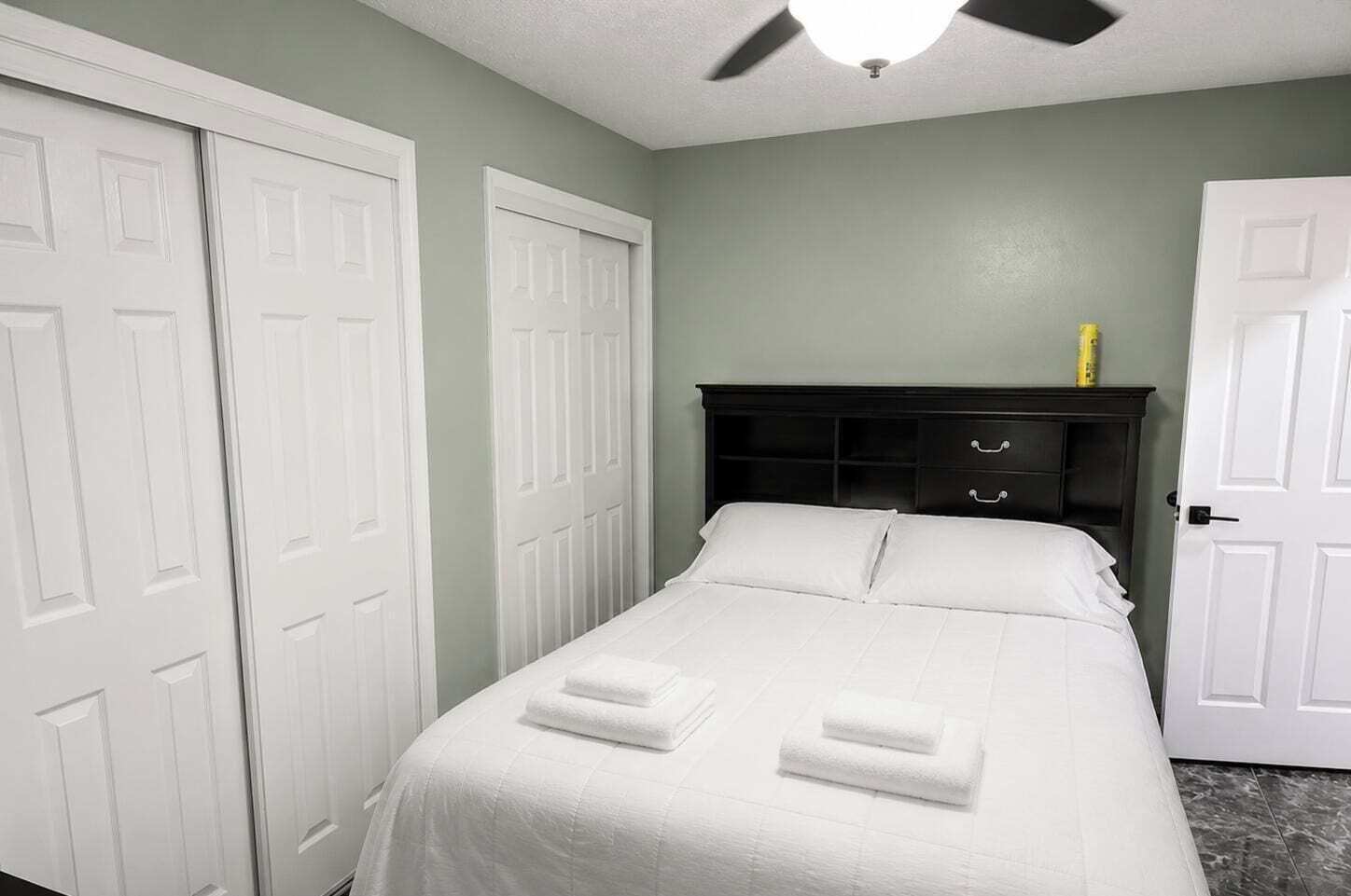 A bedroom with a bed, two white doors, a dark headboard, and a ceiling fan.
