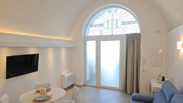 A modern living room with a flat-screen TV, a dining table with chairs, a blue sofa, and a large arched window with a decorative glass design. - SYRENIA HOME (Vico Equense)