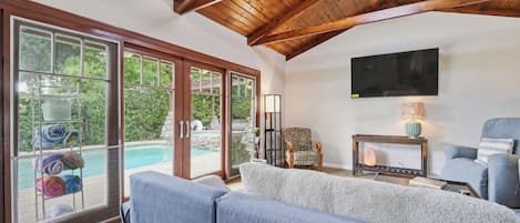 A living room with a large window, a flat-screen TV, and a pool visible through the glass doors.