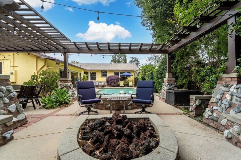 A covered outdoor patio with a fire pit, two chairs, and a pool in the background.