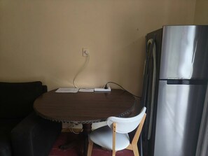 Una mesa de madera redonda con papeles, una silla blanca y un refrigerador de acero inoxidable en una habitación con una pared color beige y un enchufe.