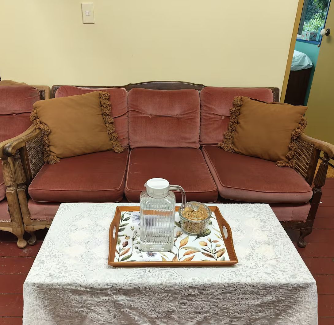 Um sofá vermelho com almofadas castanhas e uma poltrona de madeira, uma mesa branca com toalha de renda e uma bandeja com jarra de vidro e um recipiente com nozes.