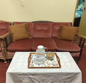 A red sofa with brown cushions and a wooden armchair, a white table with a lace tablecloth, and a tray with a glass pitcher and a bowl of nuts.