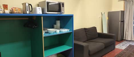 A living room with a blue shelving unit, a grey sofa, a refrigerator, and a striped curtain.