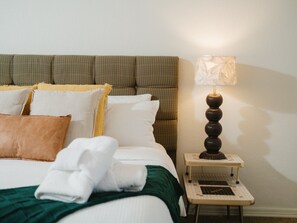 A neatly made bed with a green throw, white pillows, and a brown pillow. There is a bedside table with a lamp and a small white tray.