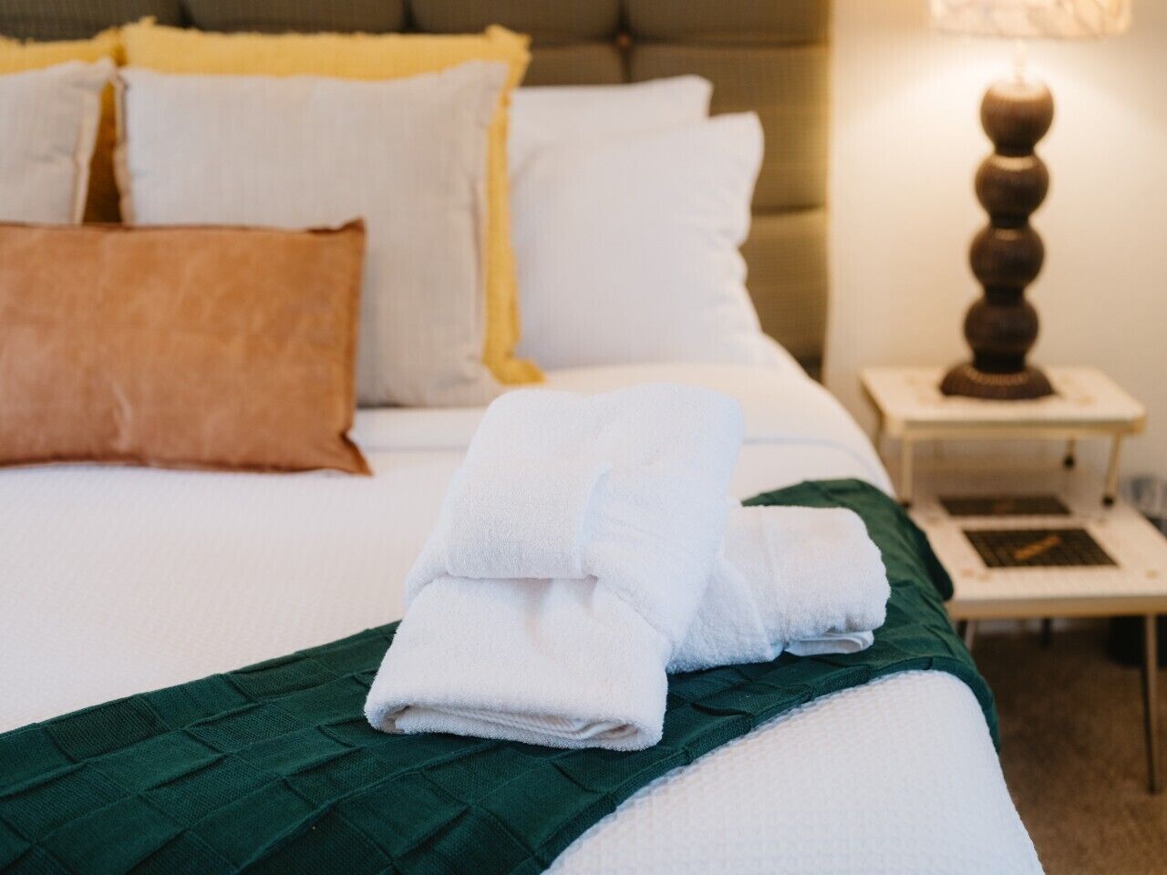 A neatly made bed with white linens and a green throw blanket, a bedside table with a lamp, and a brown pillow.