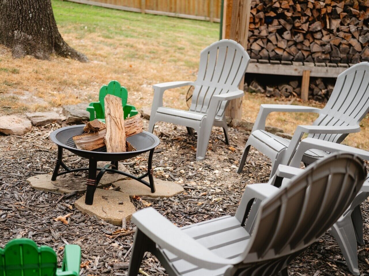 Un espacio al aire libre con sillas Adirondack, un brasero con leña y un montón de leña al fondo.
