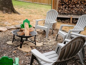 A patio area with Adirondack chairs, a fire pit with wood, and a pile of firewood in the background.