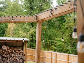 A wooden pergola with hanging light bulbs, a wooden shed, and a pile of firewood.
