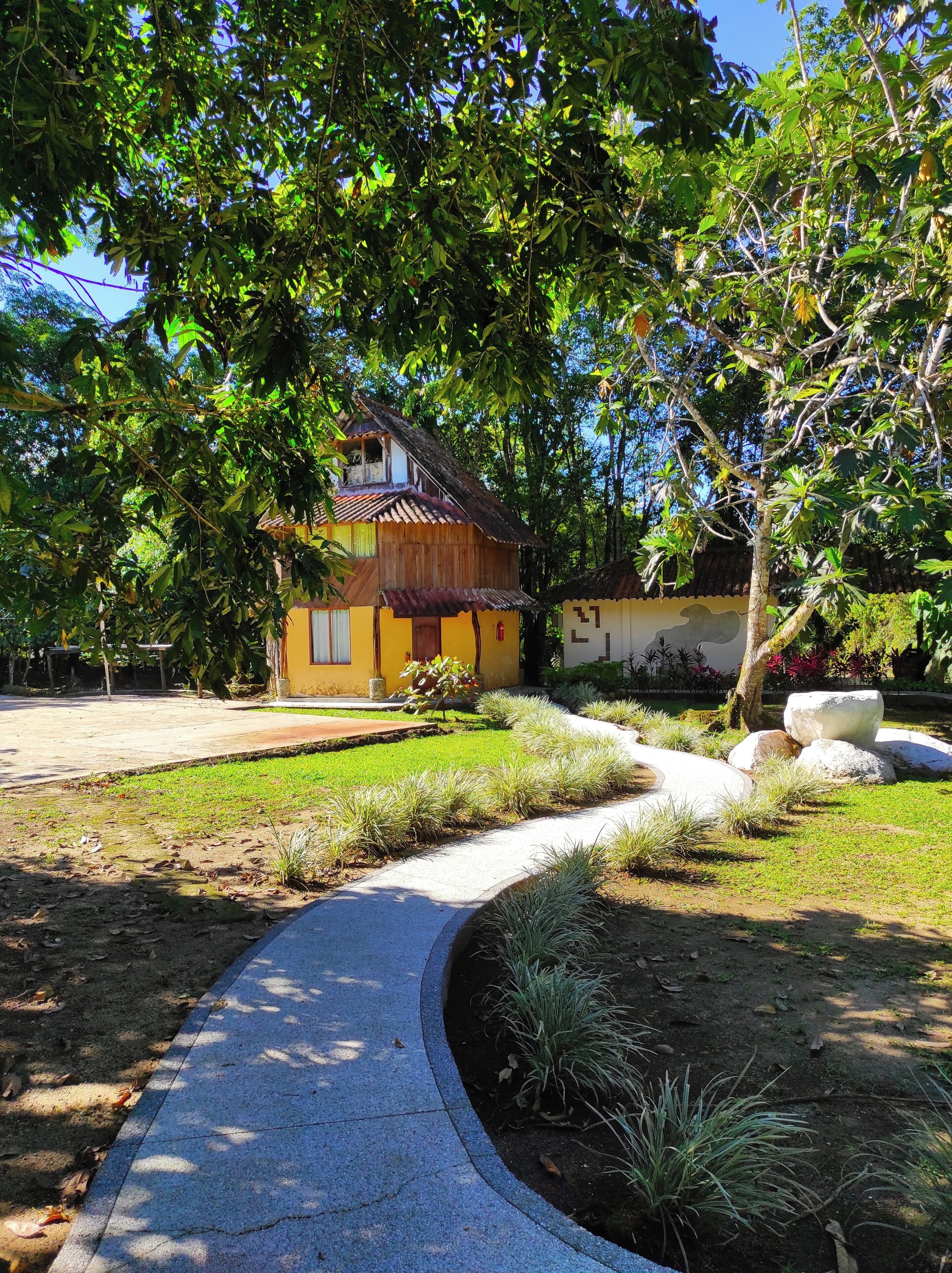 Una propiedad residencial con entrada pavimentada, un pequeño jardín con césped y arbustos, y una casa de madera con techo de tejas.