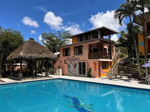 A building with a pool, a thatched-roof structure, and a staircase leading to a balcony.