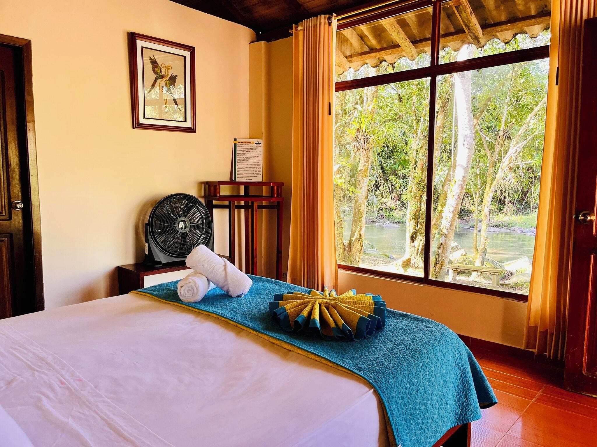A hotel room with a bed, a fan, a window with curtains, and a framed picture on the wall.